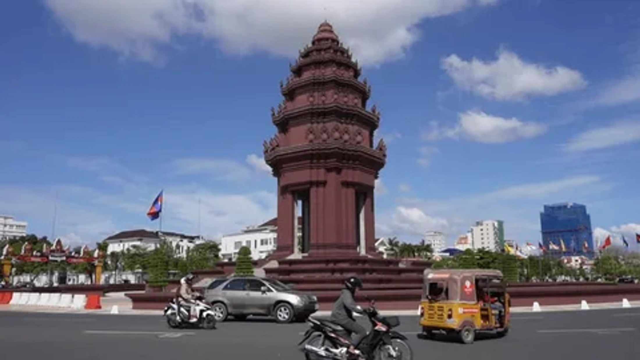 The reddish-brown Independence Monument stands in a traffic circle in Phnom Penh, with motorcycles and a tuk-tuk driving by under a blue sky.