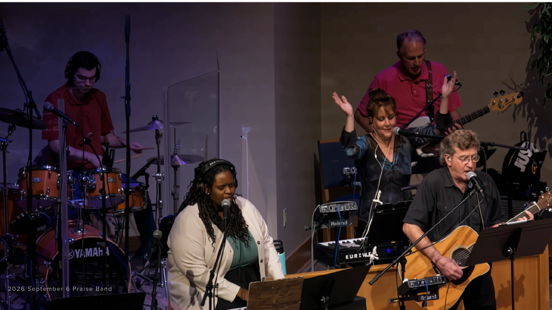 A five-member praise band performs on stage. A drummer, keyboardist/vocalist, guitarist/vocalist, another vocalist, and a bassist play their instruments.