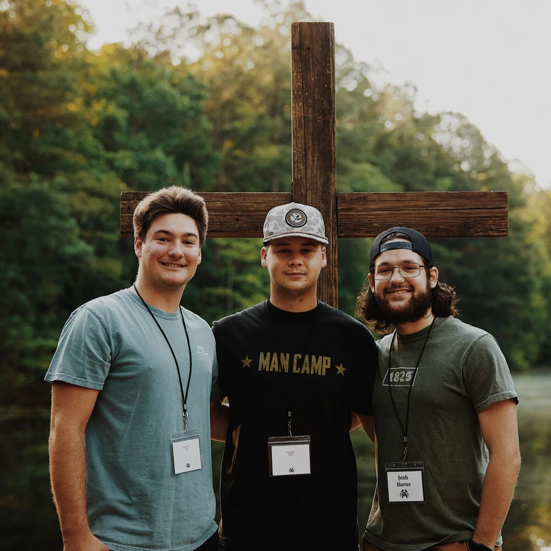 Three young men smile for a photo while standing in front of a large wooden cross outdoors. They are wearing lanyards and casual clothes at a camp.