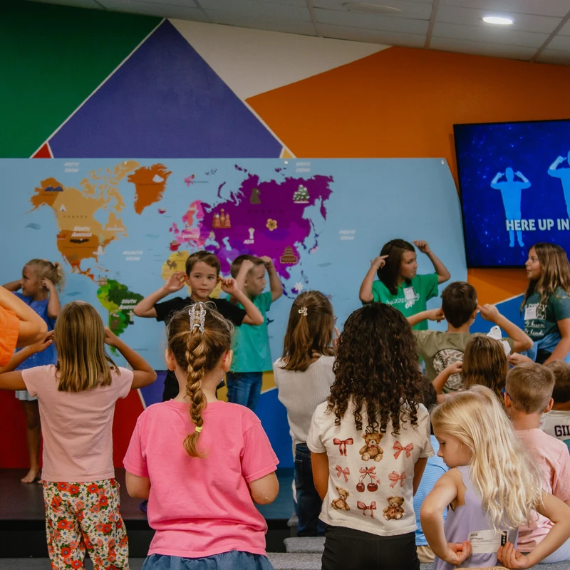 A group of children in a colorful classroom follow along with a song, mimicking actions shown on a screen in front of a large world map.