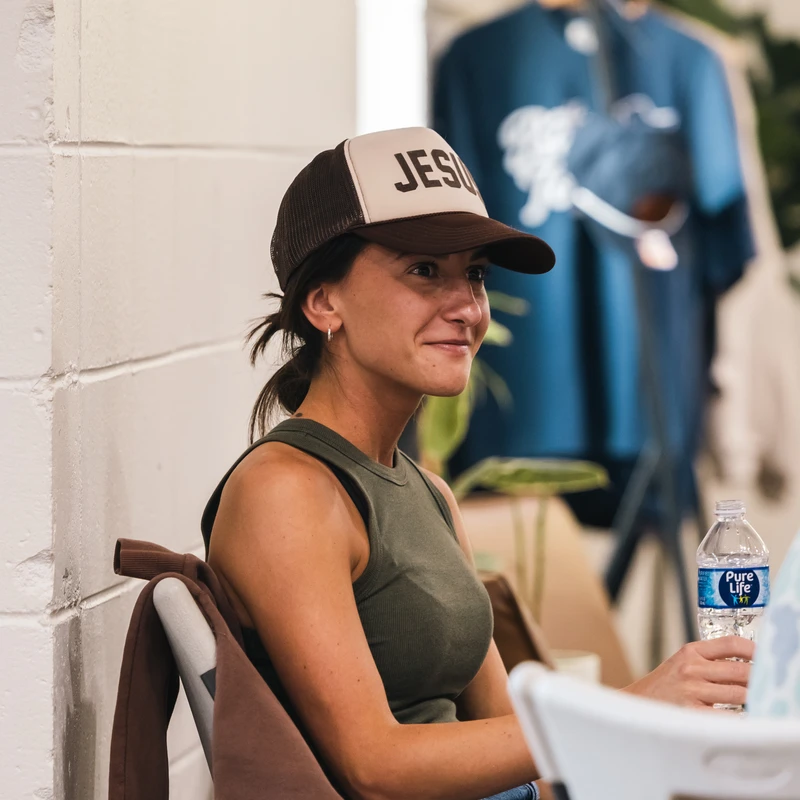 A smiling woman with dark hair sits indoors, wearing a brown and beige trucker hat with 'JESUS' on the front, and holding a bottle of water.