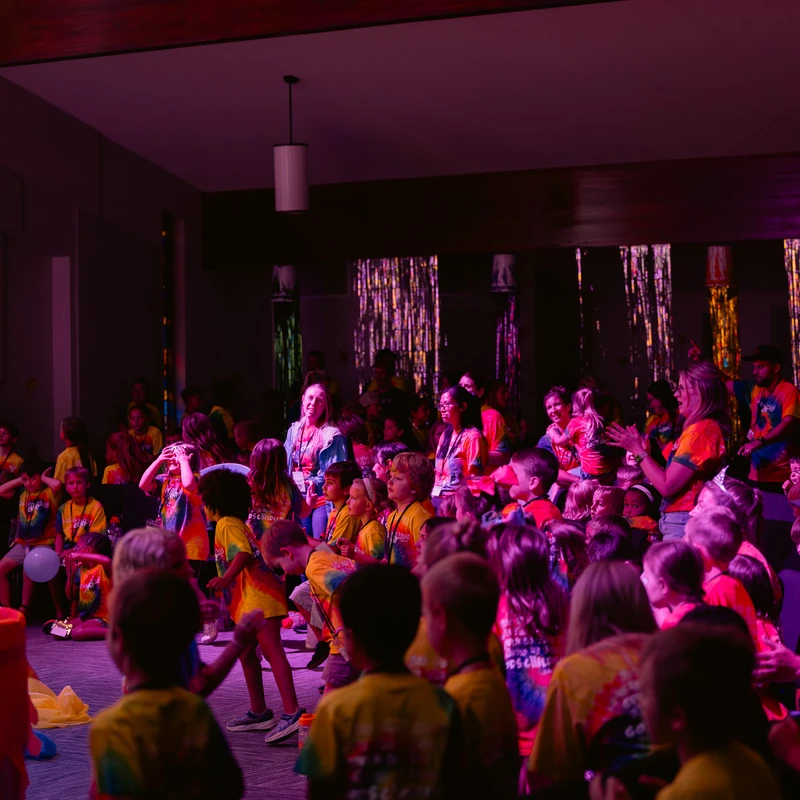 A large crowd of children and adults in colorful shirts watch a performance in a large room lit with pink and purple lights during a summer camp assembly.