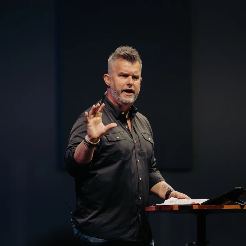 A man with short gray hair and a beard in a black shirt stands at a lectern, speaking and gesturing with his left hand. An open book rests on the lectern.
