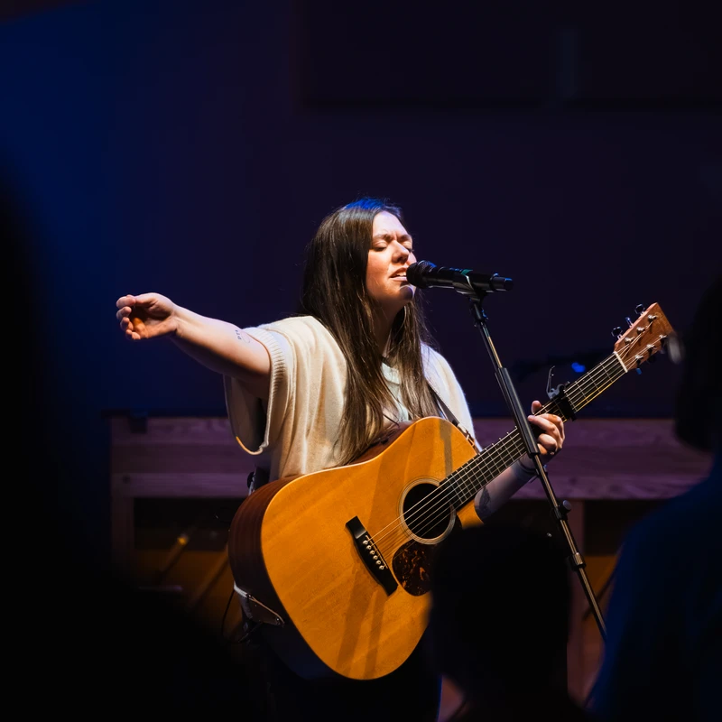 A woman with long dark hair sings passionately into a microphone while playing an acoustic guitar on a dimly lit stage, her arm outstretched.