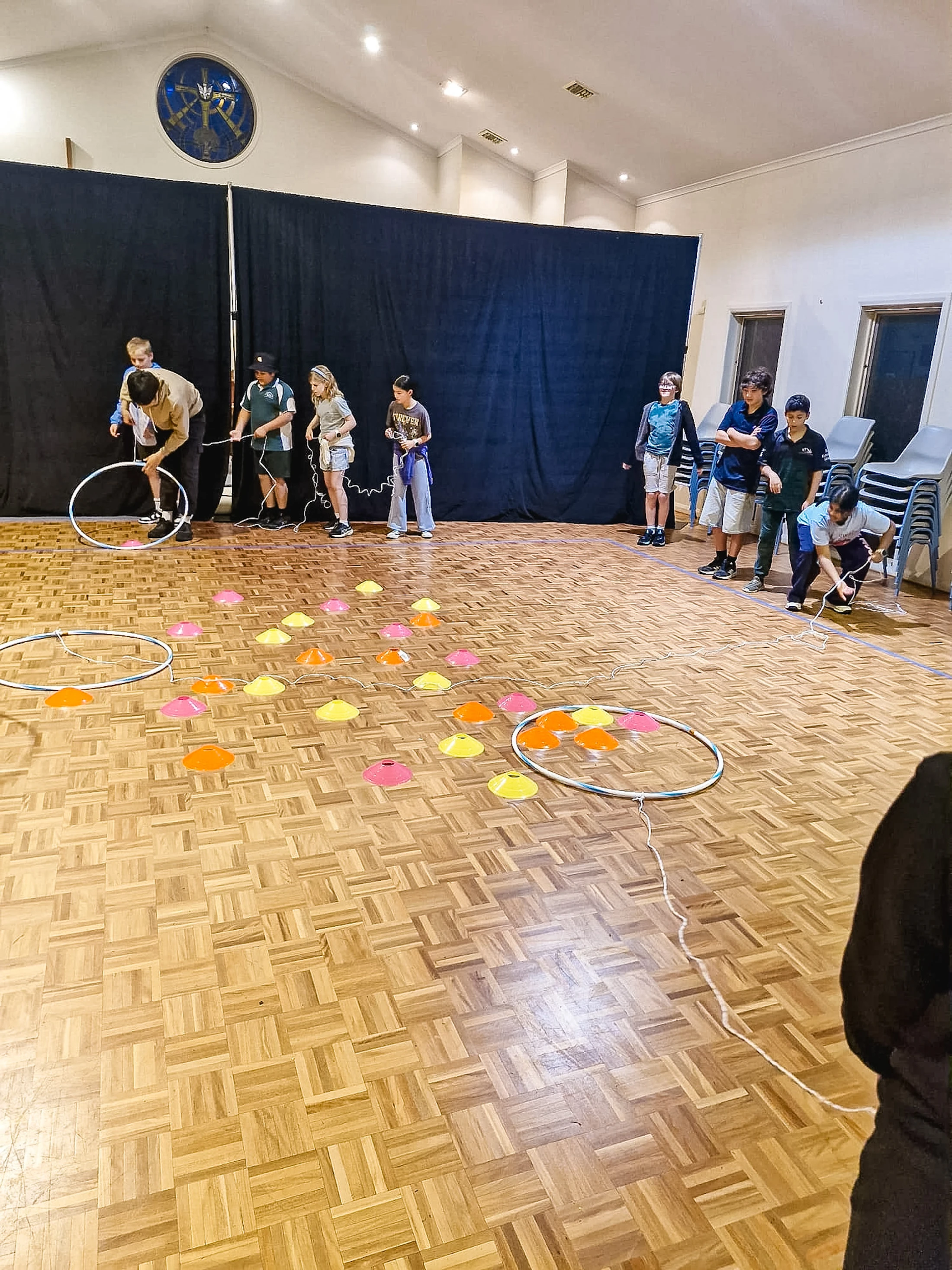 A group of children and adults participate in a team game inside a large hall, using hula hoops, ropes, and colorful cones on a wooden parquet floor.