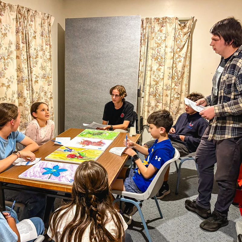 A group of children and teenagers sit around a table with their paintings, listening as a young man in a plaid shirt stands and addresses them.