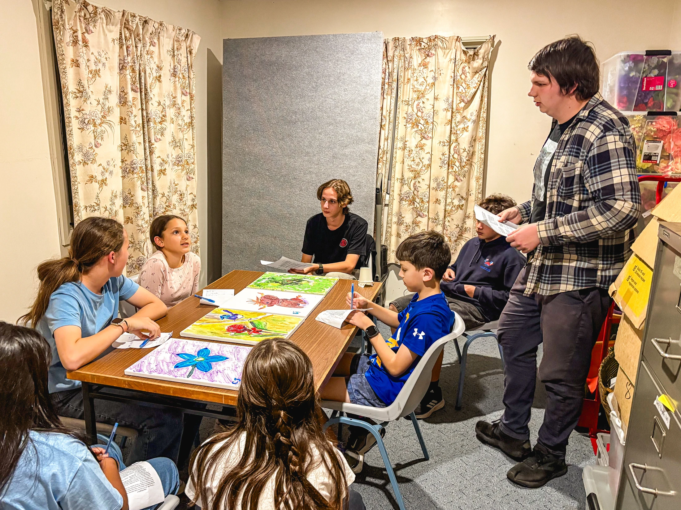 A group of children and teenagers sit around a table with their artwork while a young man stands and leads a discussion in a classroom.