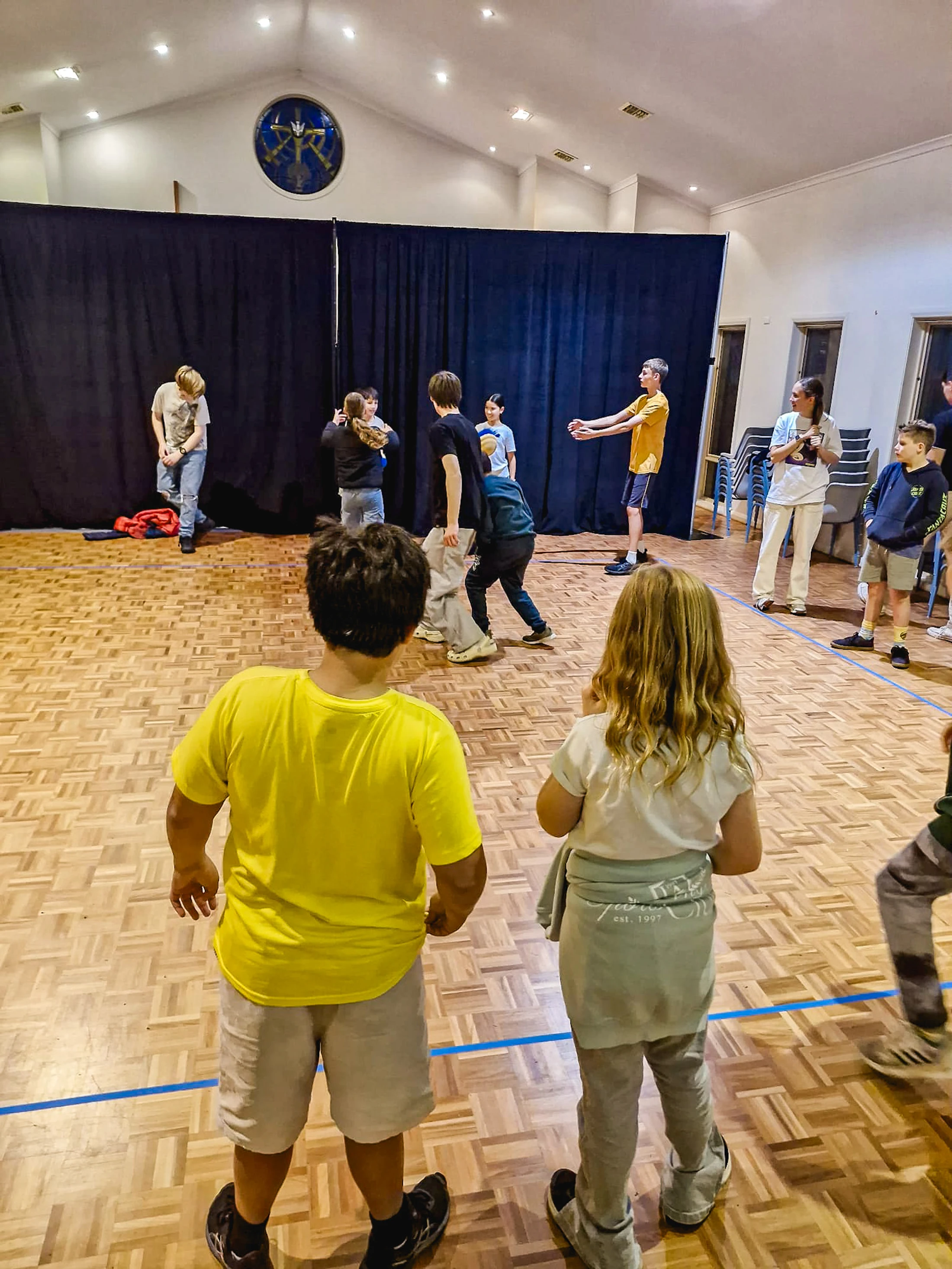 A group of children and teenagers rehearsing on a parquet floor in a large hall, with a black curtain backdrop and two kids watching from the foreground.