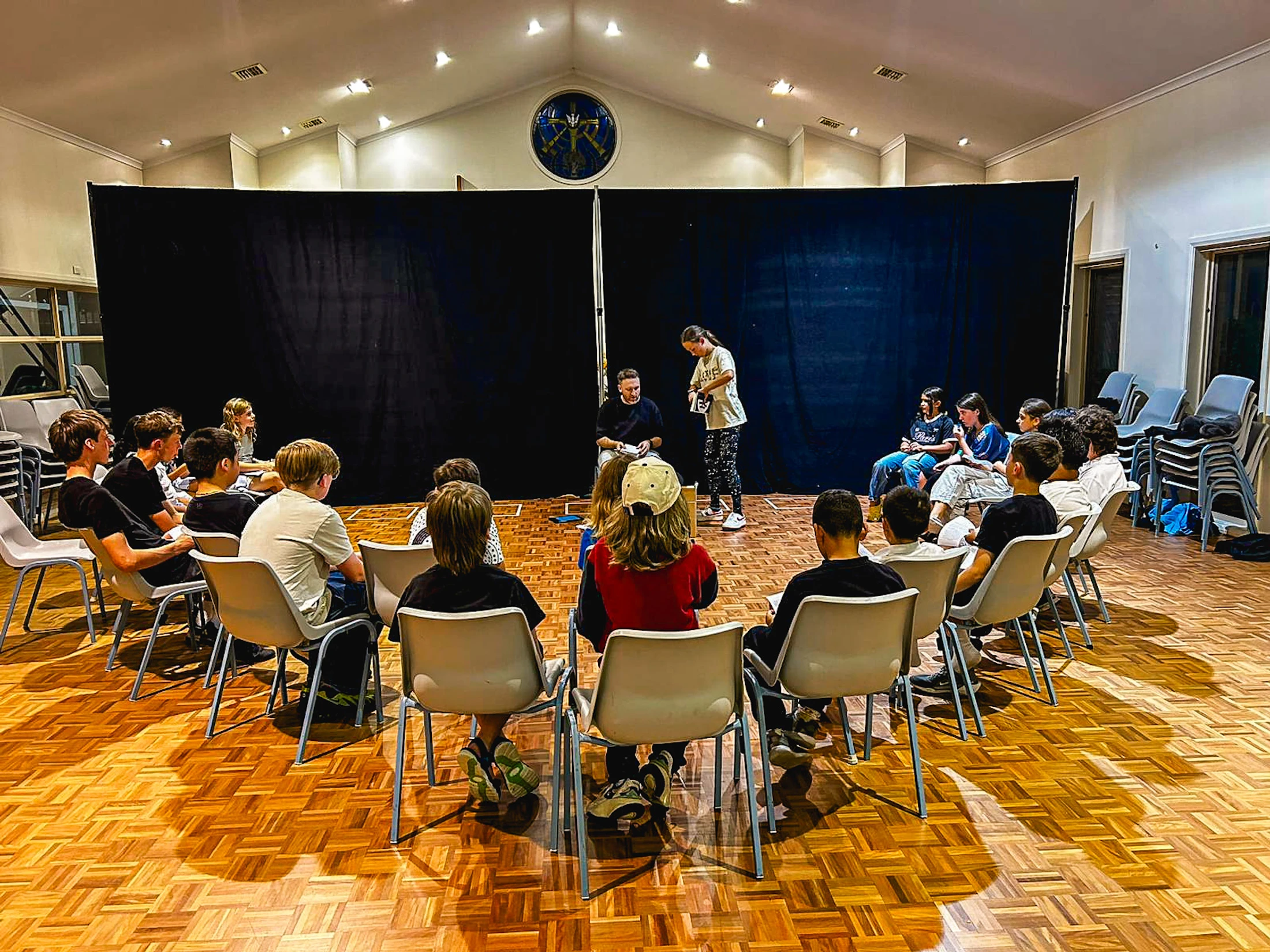 A group of young people sit in a semi-circle on chairs, watching two adults in front of a black backdrop during a workshop in a large hall.