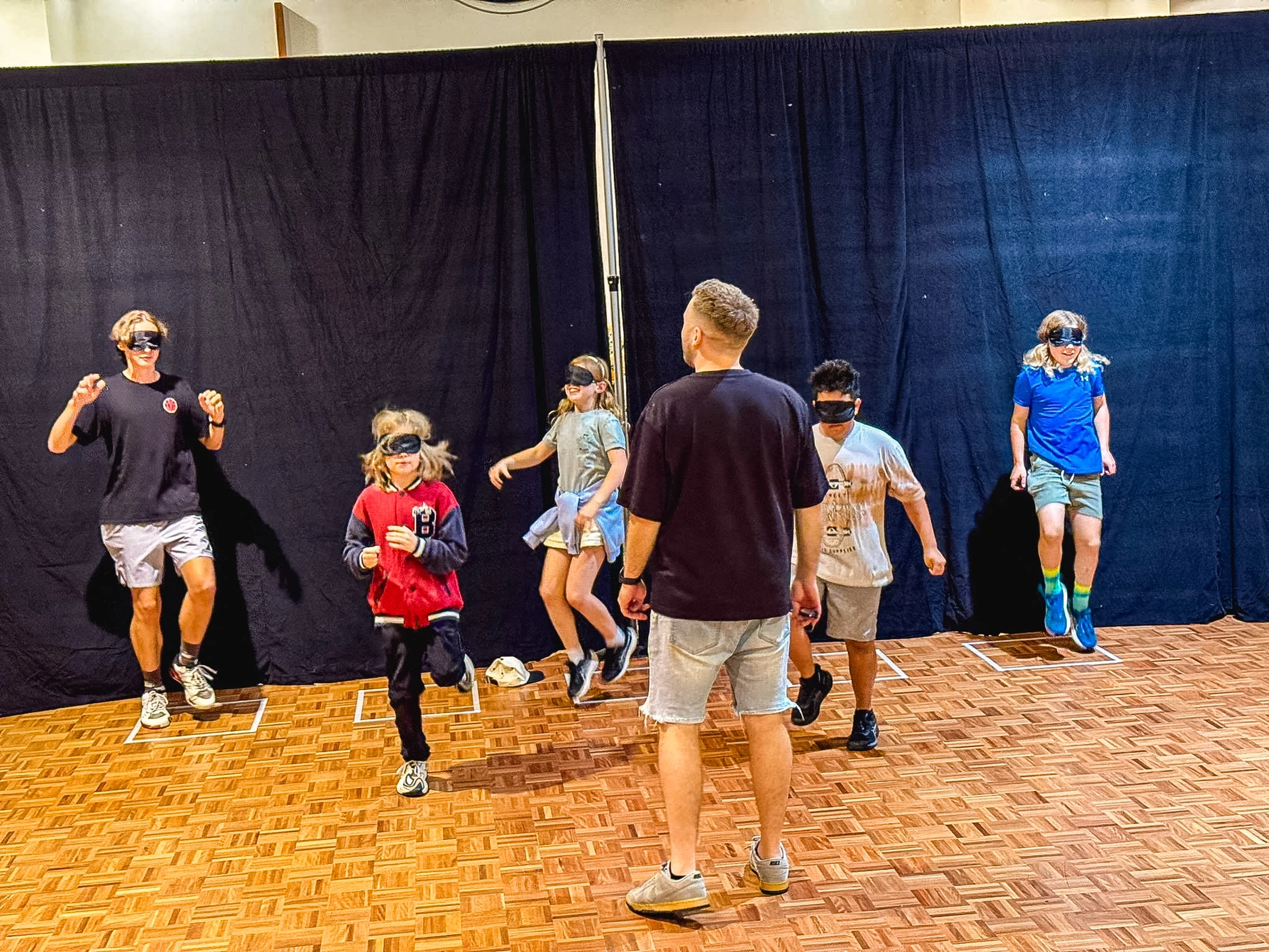 A group of blindfolded children run and jump on a wooden floor, guided by an adult during an activity in front of a black curtain.