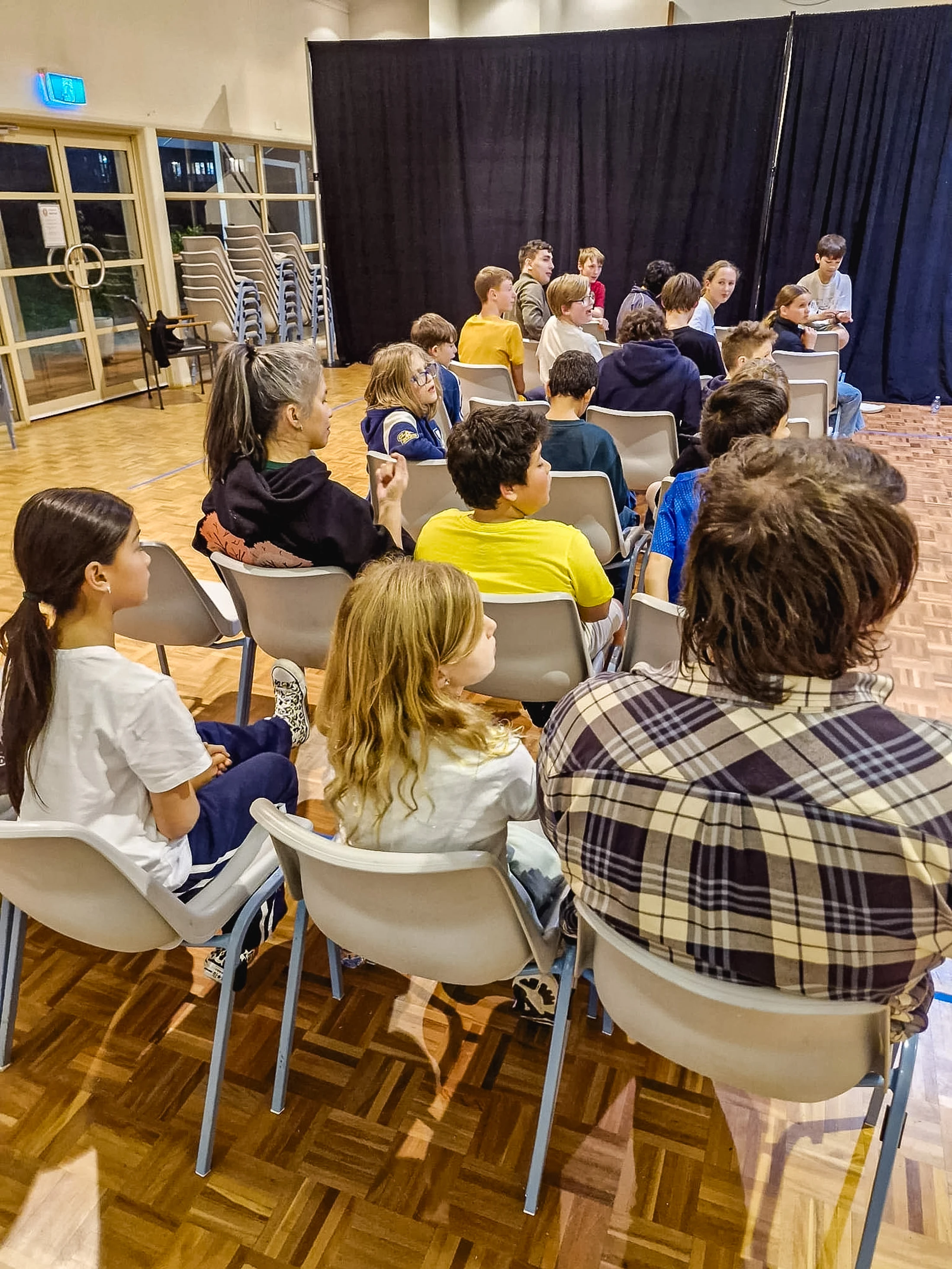 A group of young people sit on chairs in a hall with a wood floor. They are viewed from behind as they watch a presentation in front of a black curtain.