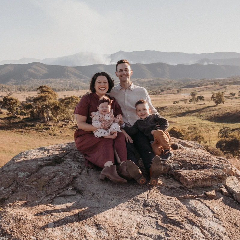 A smiling family of four, parents with a young son and baby daughter, sit together on a large rock with rolling hills and mountains in the background.