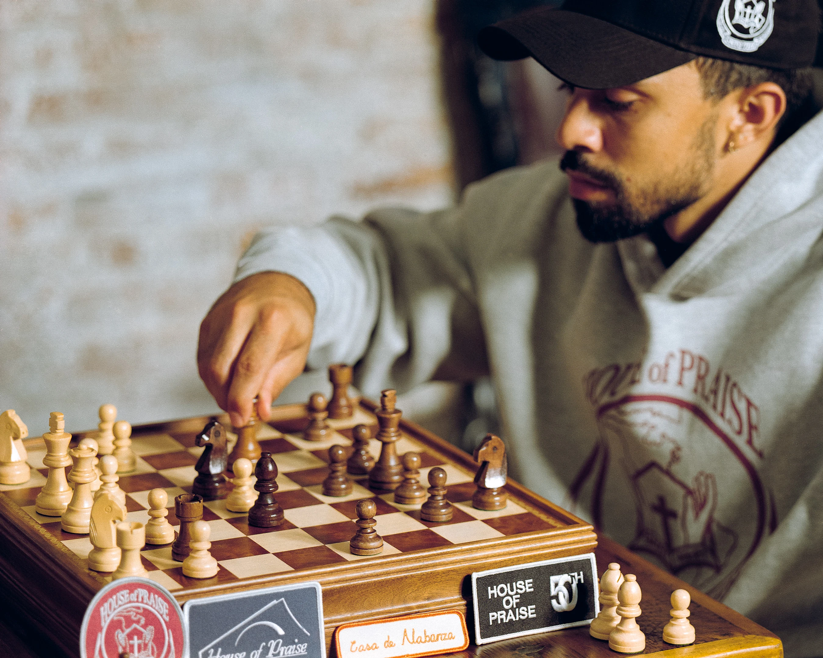 A man with a beard, wearing a grey hoodie and a black cap, leans over a wooden chessboard and moves a piece, focused intently on the game.