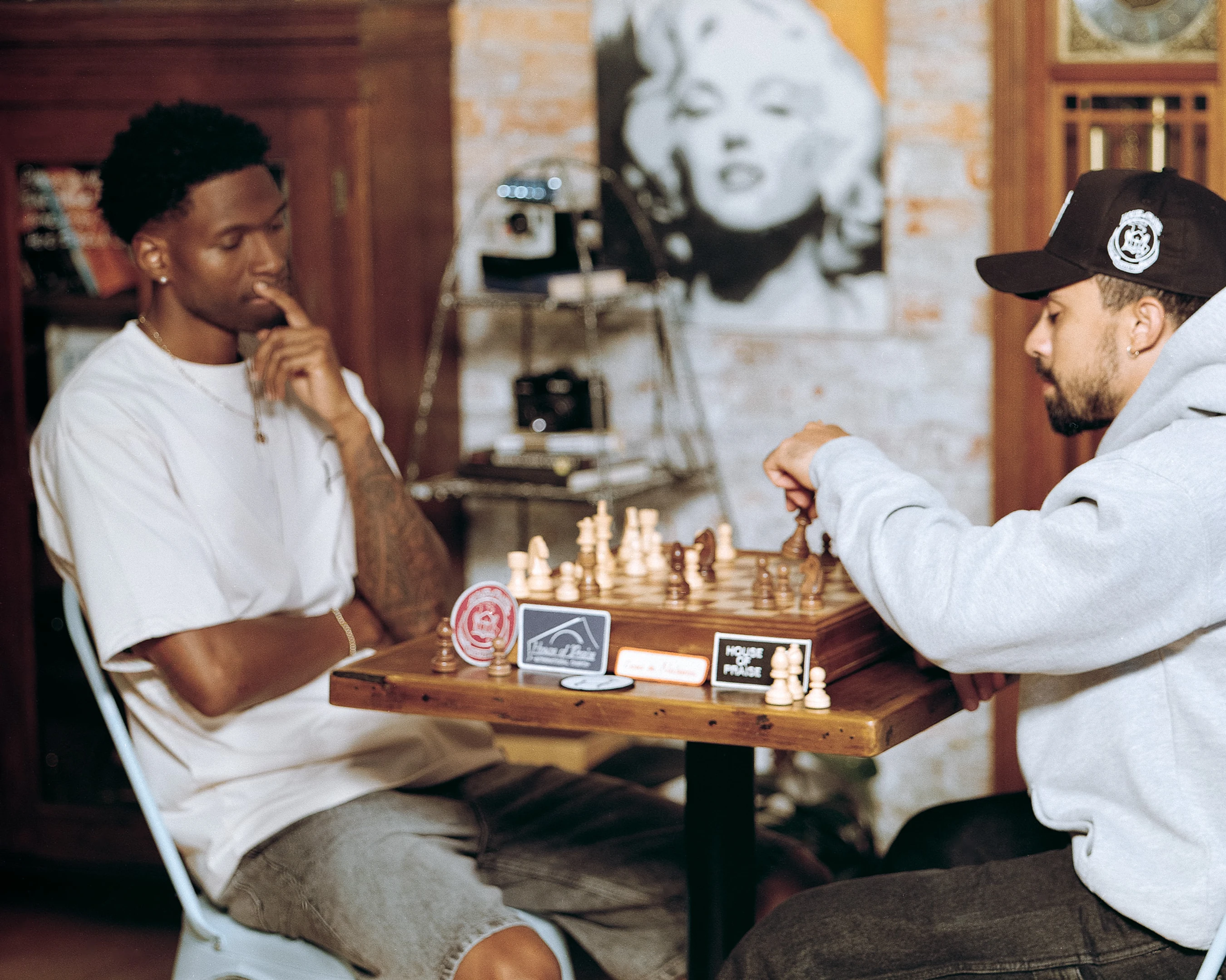 Two men sit at a small wooden table playing chess. One man in a white t-shirt thinks about his next move, while the other in a grey hoodie makes his.