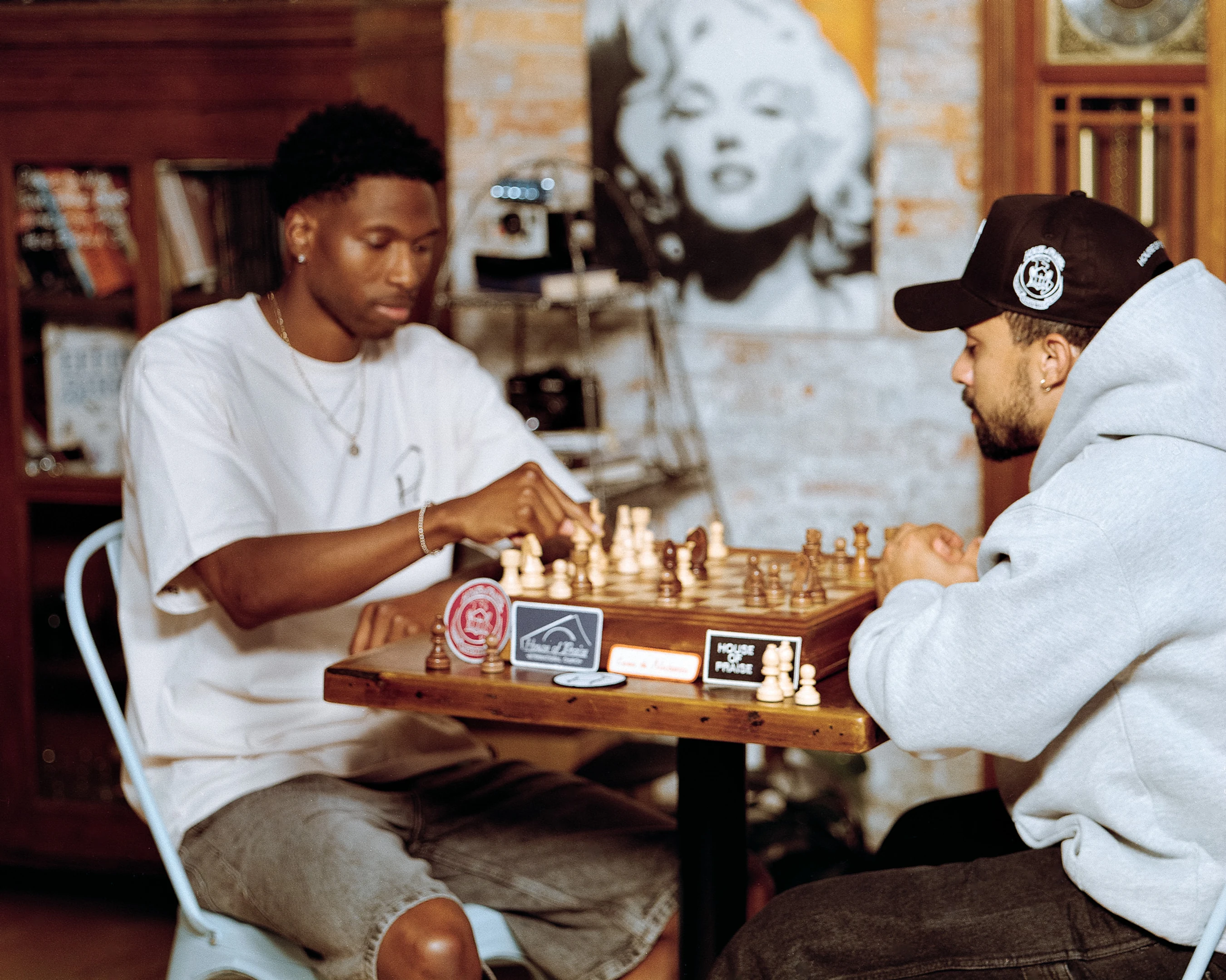 Two young men sit opposite each other at a wooden table, engaged in a game of chess. One man in a white t-shirt is making a move on the board.