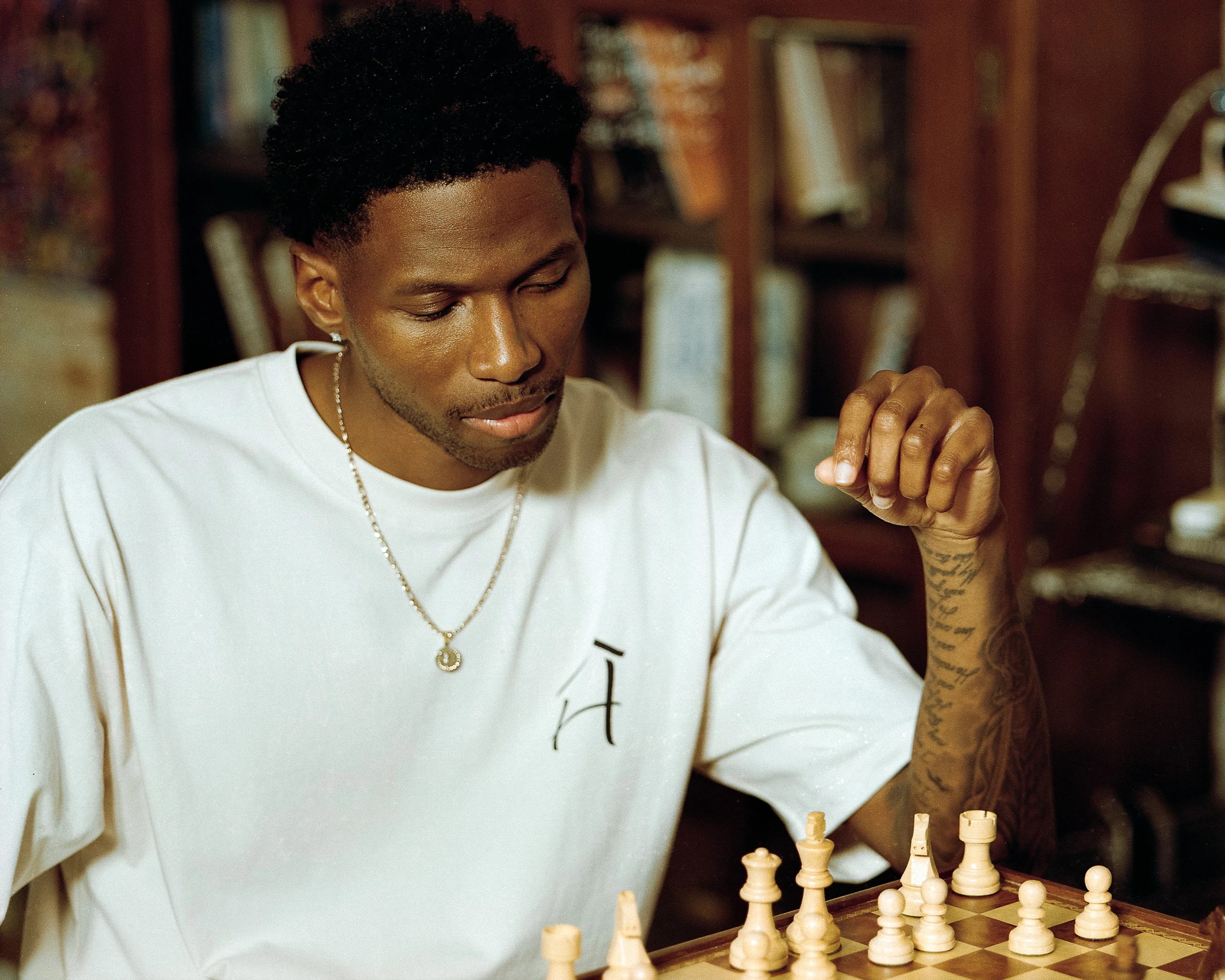 A young Black man in a white t-shirt with tattoos on his arm, looking down thoughtfully at a chessboard as he contemplates his next move.