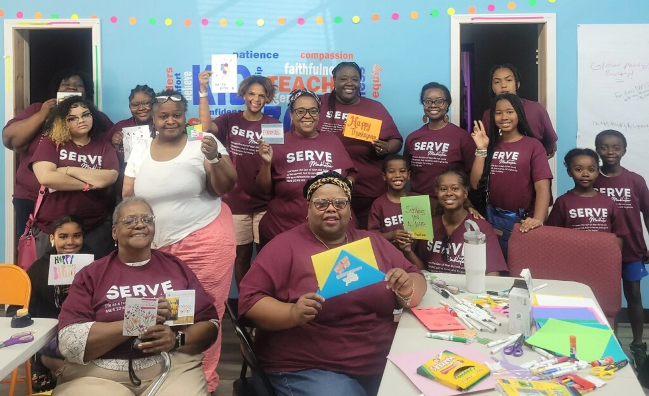 A large, diverse group of smiling volunteers wearing matching maroon 'SERVE' t-shirts pose together holding up handmade cards in a colorful classroom.
