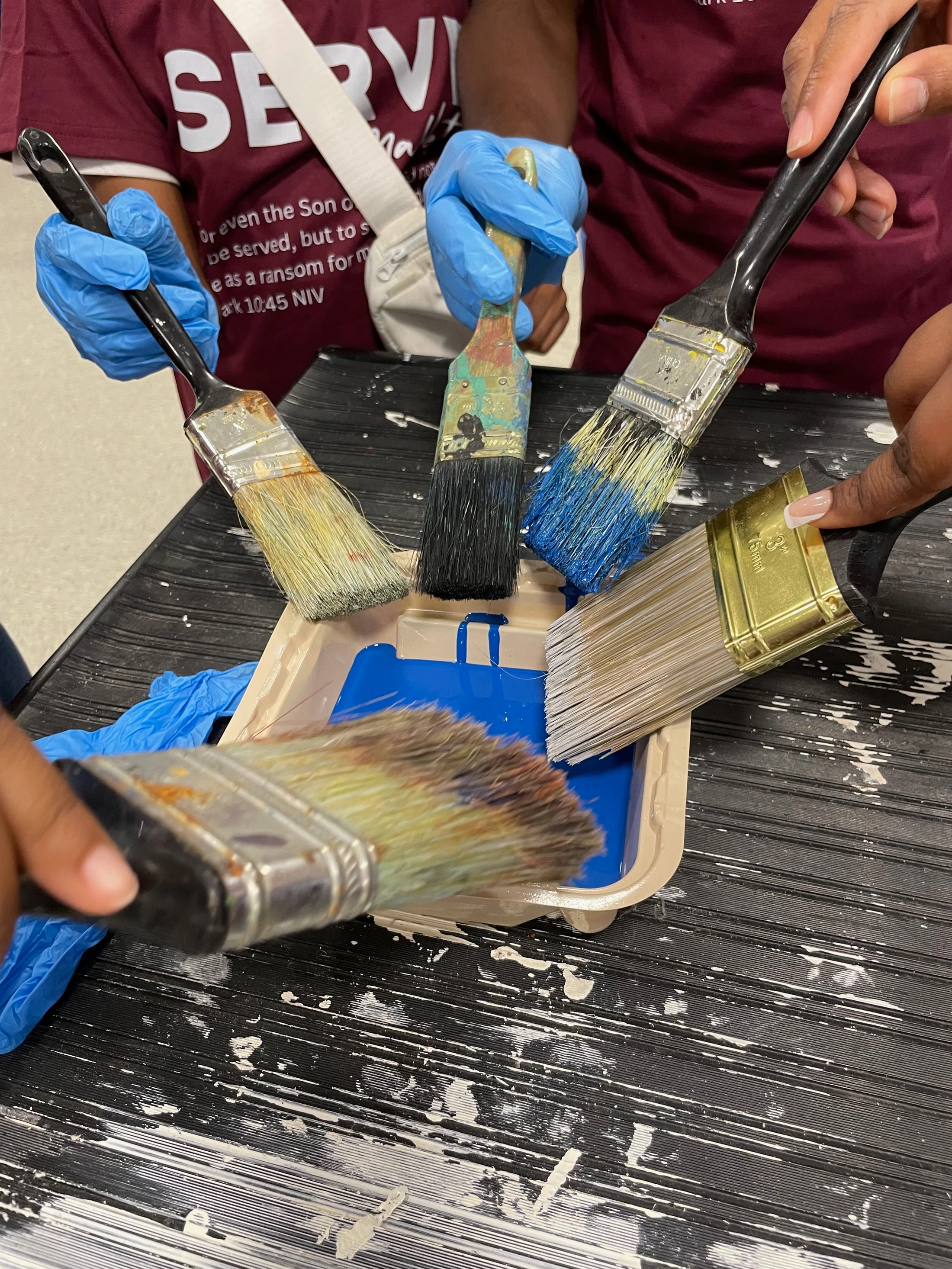 Several hands, some in blue gloves, dip paintbrushes together into a tray of bright blue paint over a paint-splattered black surface.