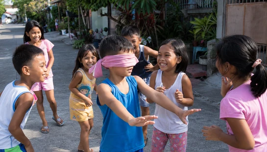 A group of happy children play a game on a paved street. One boy is blindfolded with a pink cloth and reaches out as his friends laugh and dodge him.