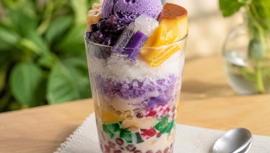 A tall glass of halo-halo with layers of sweet beans, jellies, shaved ice, and milk, topped with a scoop of ube ice cream and a slice of leche flan.
