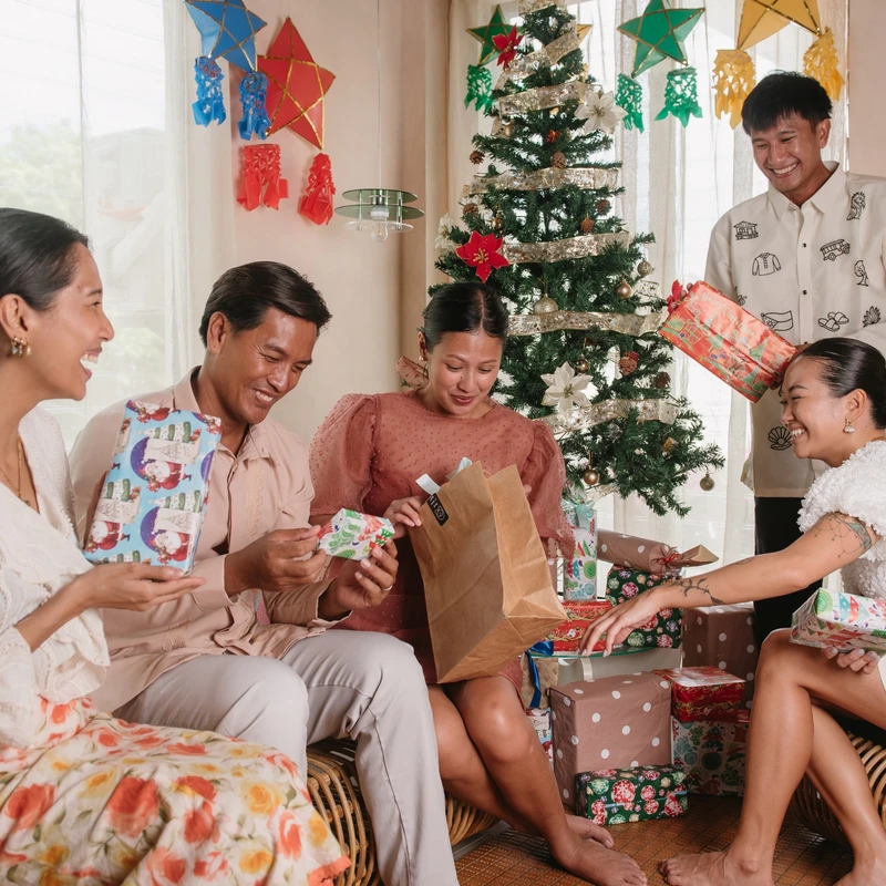 A family of five smiling Filipino adults exchange wrapped gifts in a living room decorated with a Christmas tree and traditional star-shaped lanterns.
