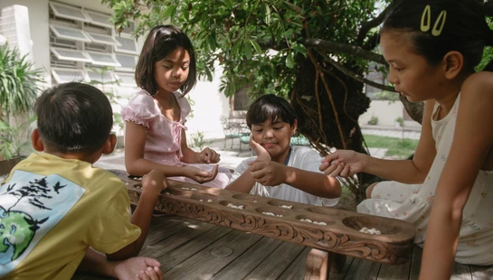 Four children gather around a wooden table in a yard, intently playing a traditional carved wooden board game, known as Sungka or Mancala.