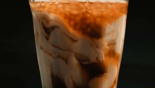 A close-up shot of a glass filled with taho, showing layers of soft white tofu, dark brown arnibal syrup, and sago pearls against a dark background.