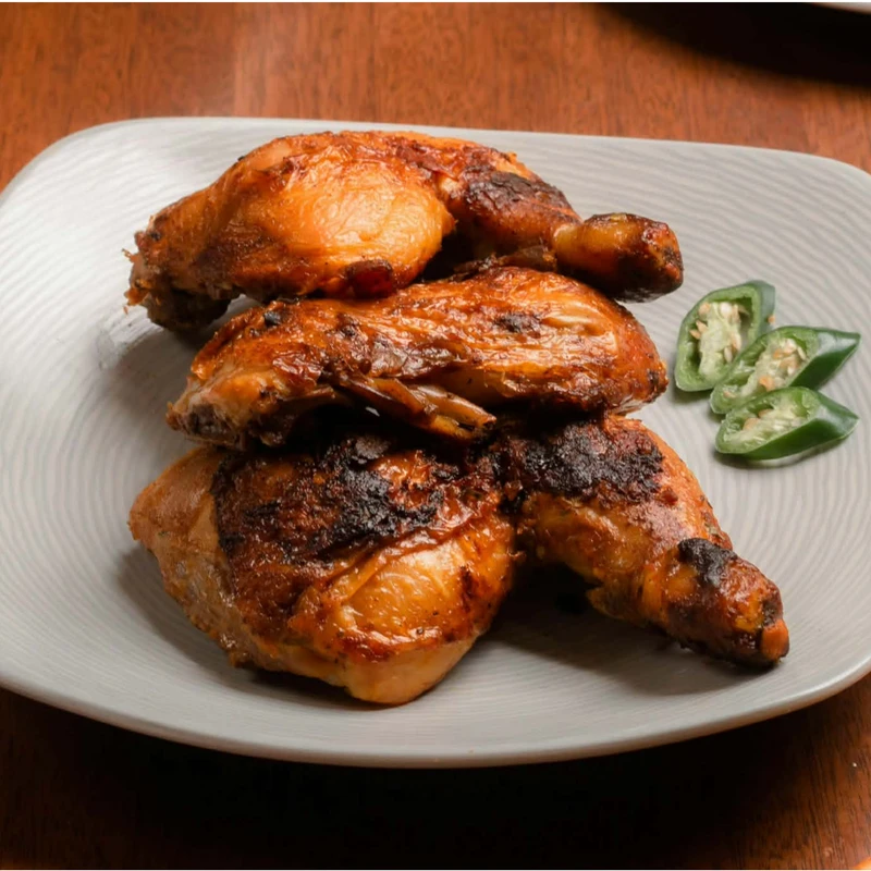 Three crispy, golden-brown roasted chicken leg quarters stacked on a light grey plate, garnished with slices of fresh jalapeño, on a wooden table.