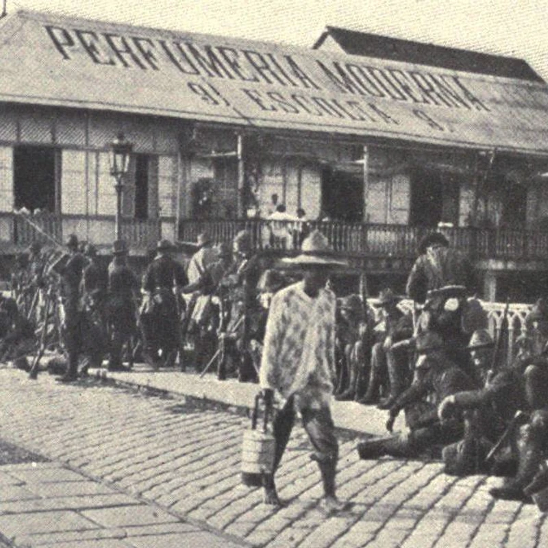 A black and white historical photo of American soldiers resting on a cobblestone street in Manila. A Filipino man walks past them in front of a building.