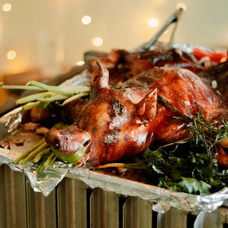 A whole roasted pig with crispy skin on a foil platter, garnished with green herbs and vegetables, with warm bokeh lights in the background.