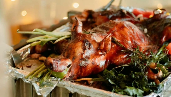 A whole roasted pig with crispy skin on a foil platter, garnished with green herbs and vegetables, with warm bokeh lights in the background.