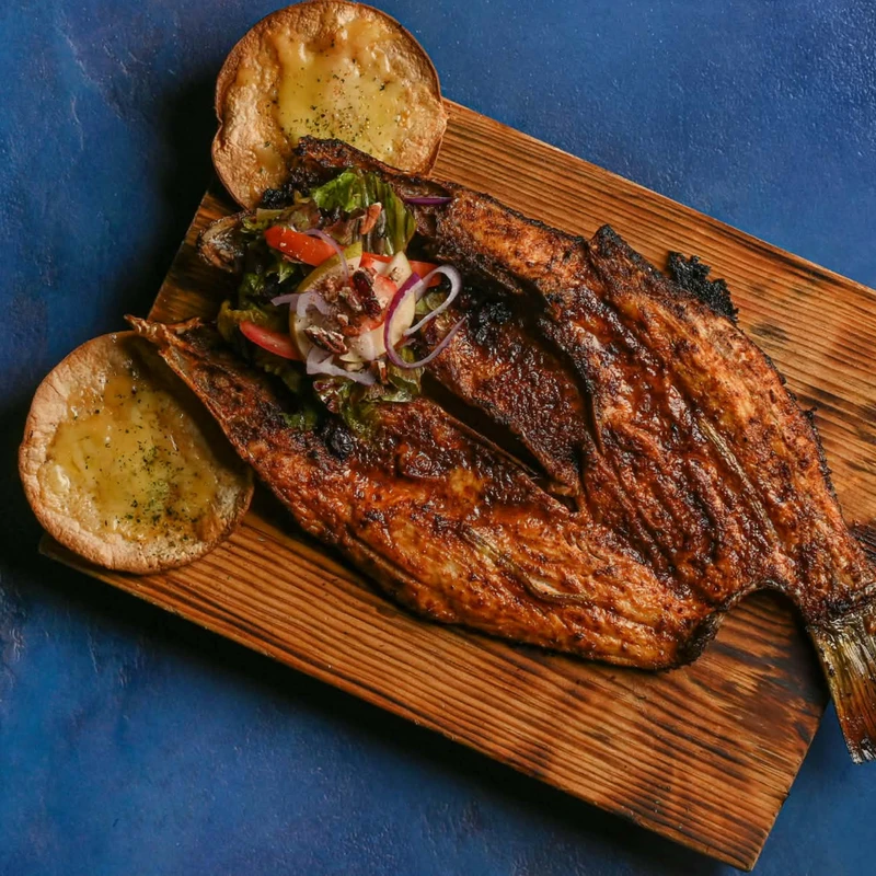 A whole grilled fish, split open, served on a rustic wooden plank with a fresh salad and two cheesy toasted flatbreads on a blue background.