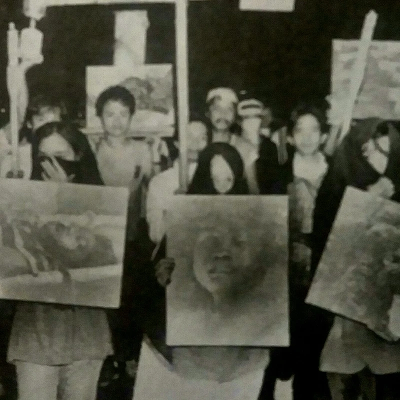A grainy, black and white photo of a group of people, mostly women, marching at night. They hold up signs with portraits, likely of victims or missing persons.