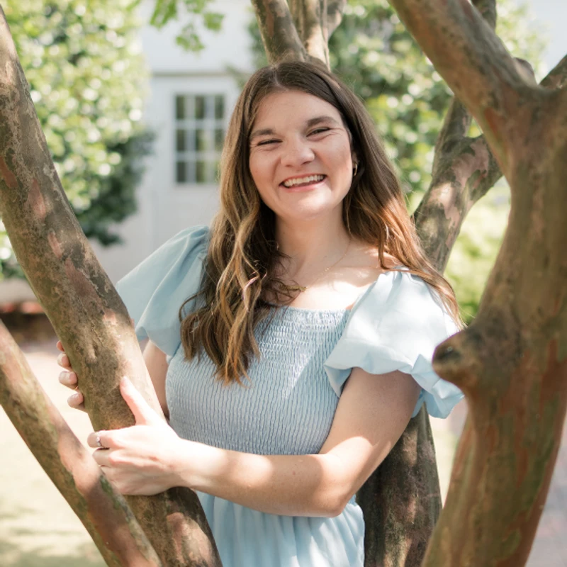 A smiling young woman with long brown hair, wearing a light blue dress, poses for a portrait while standing among the branches of a tree in a sunny garden.