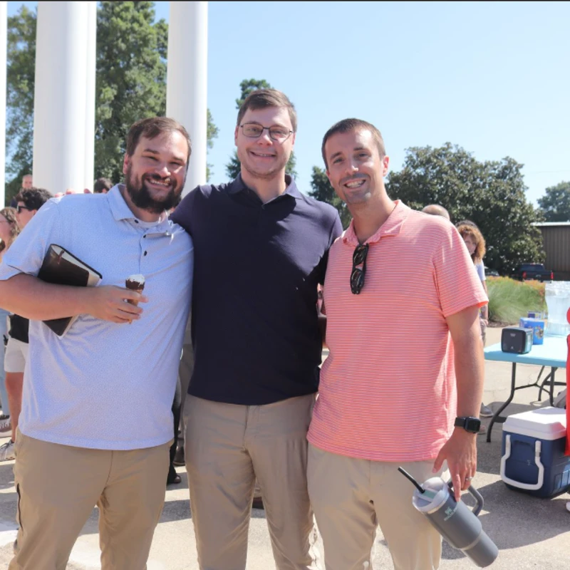 Three men stand with their arms around each other, smiling for a photo outdoors on a sunny day in front of a building with large white columns.