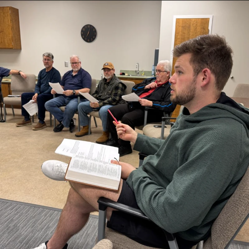 A young man in a green hoodie speaks while holding a book, as a group of older men sit in a circle and listen during a study session in a classroom.