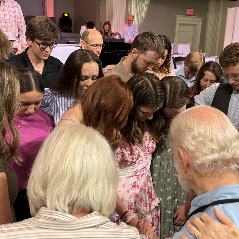 A large, diverse group of people stand in a close circle indoors, bowing their heads in prayer. Some rest hands on the shoulders of others in support.