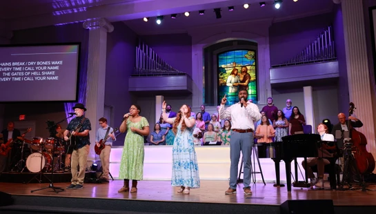 A diverse group of singers and musicians perform on a purple-lit stage in a church, with a choir and a large stained-glass window in the background.