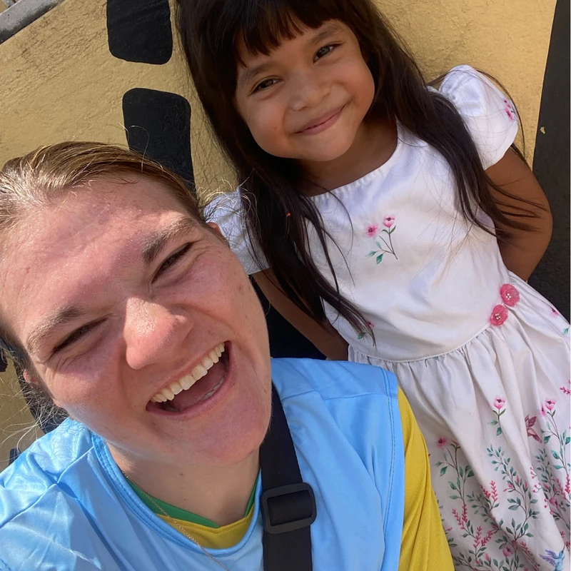 A woman in a blue and yellow top takes a joyful selfie with a young girl in a floral dress who smiles gently behind her against a yellow wall.