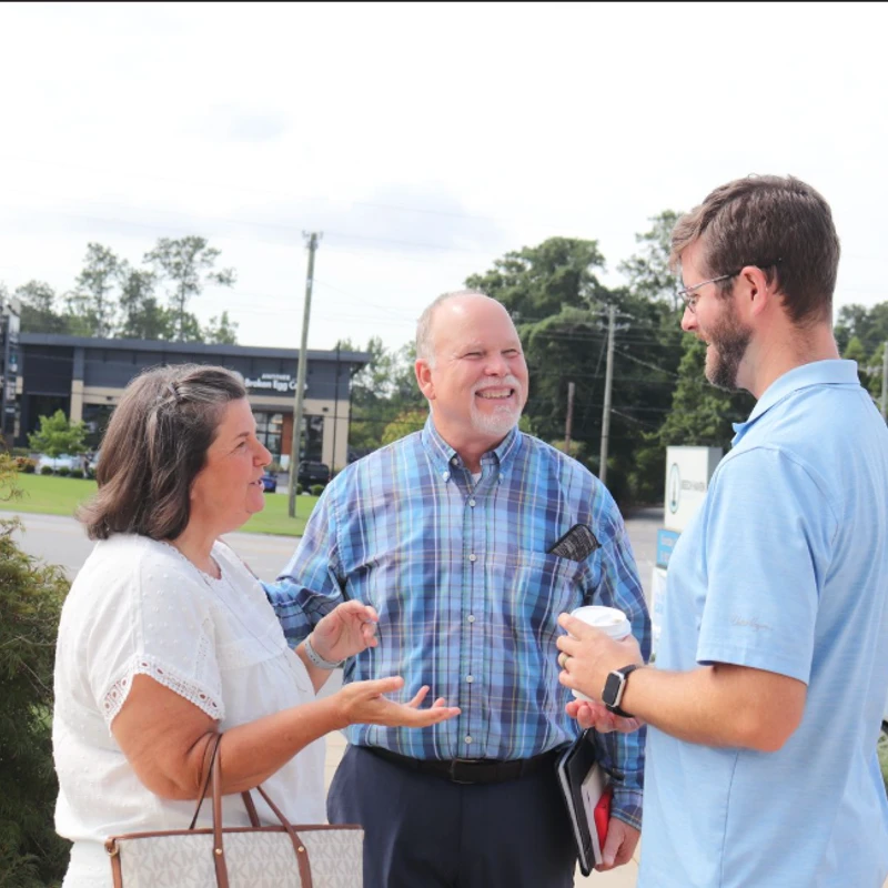 A woman in a white blouse and two men in casual shirts stand outside on a sunny day, smiling and engaged in a friendly conversation.