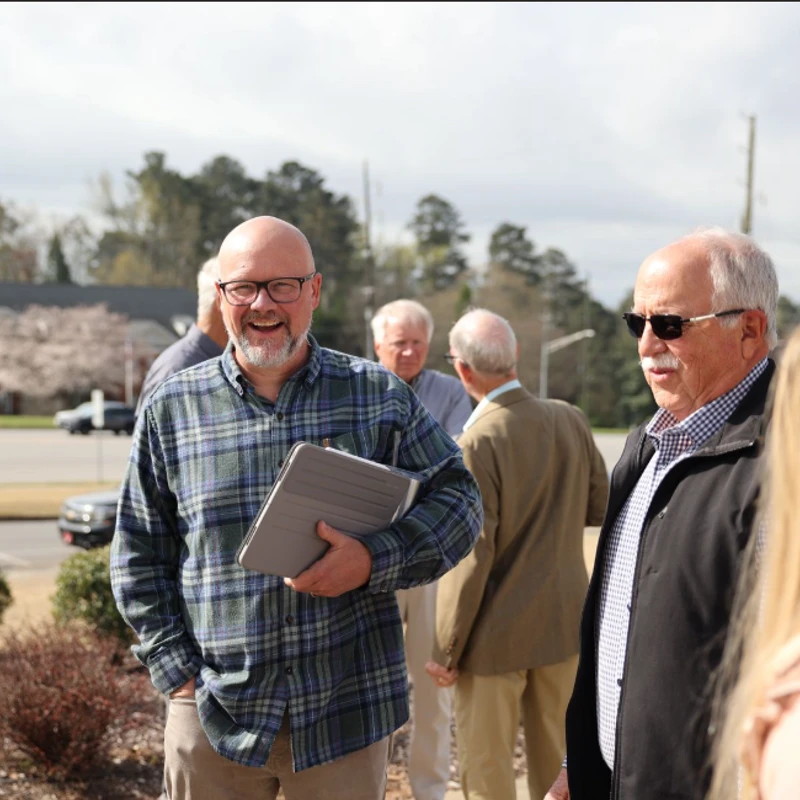 A smiling bald man with glasses and a plaid shirt holds a tablet while standing outside with a group of other men on a bright, sunny day.