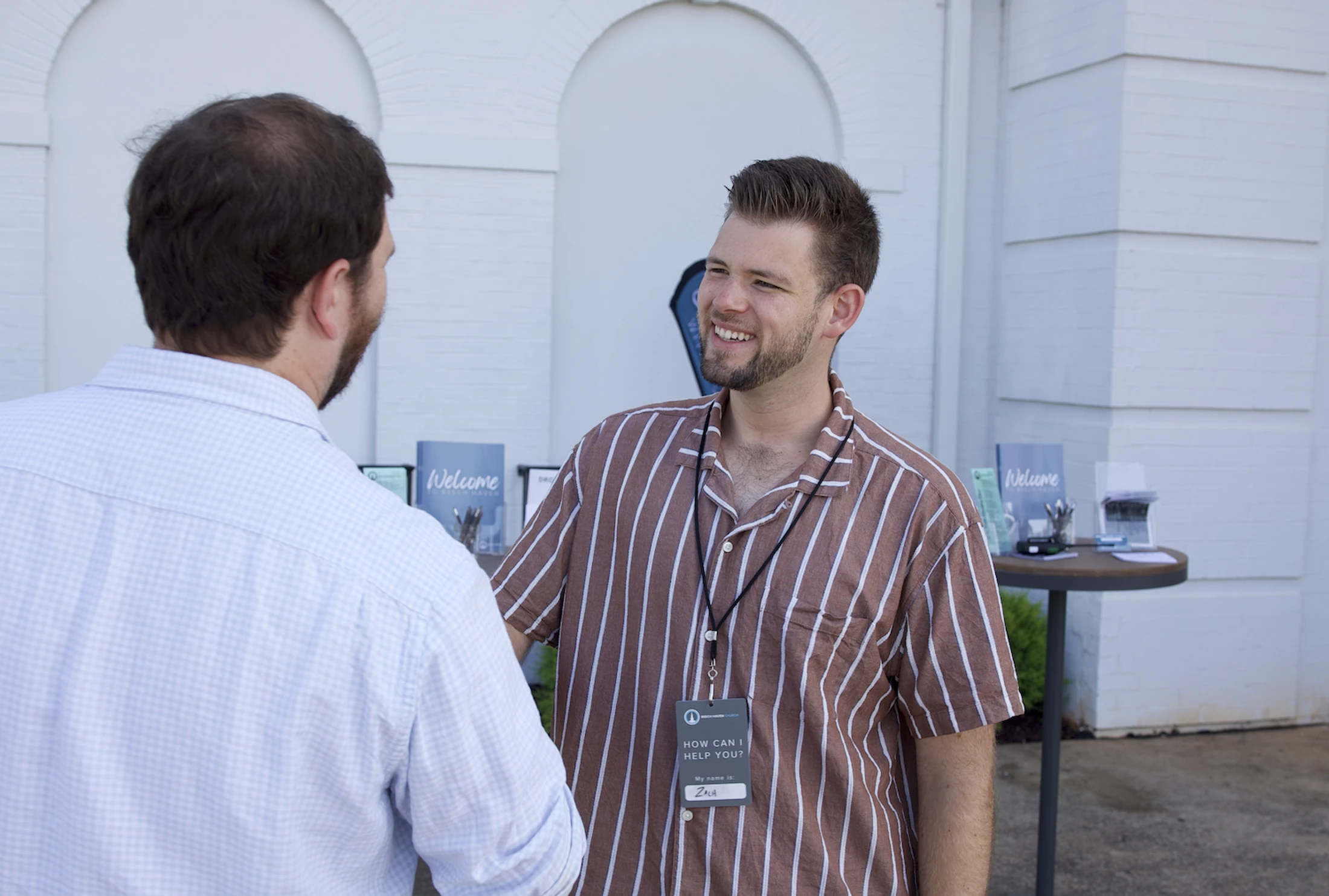 A smiling man named Zach wearing a striped shirt and a name tag greets another man outside a white building. A welcome table is in the background.