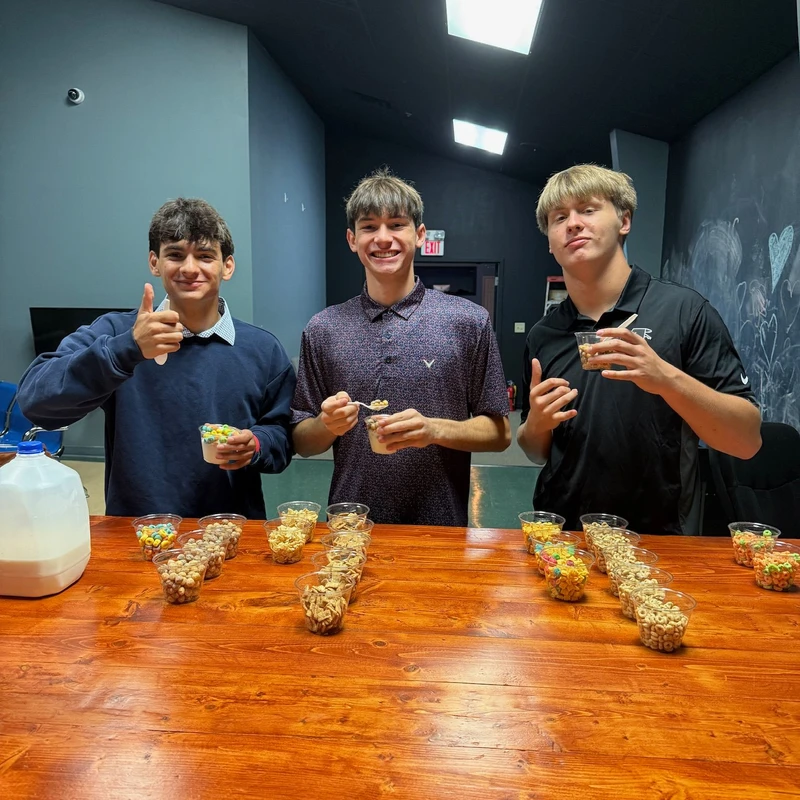 Three smiling teenage boys stand behind a wooden counter with rows of cups filled with different cereals, holding their own cups and giving thumbs up.