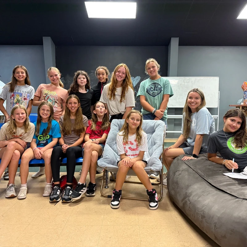 A group of thirteen young girls smiling for a photo in a casual room. They are standing and sitting on a blue bench, a soft chair, and a large beanbag.