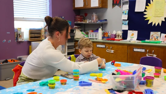 A woman and a young boy sit at a table covered in colorful modeling clay and tools, engaged in a creative arts and crafts activity in a classroom setting.