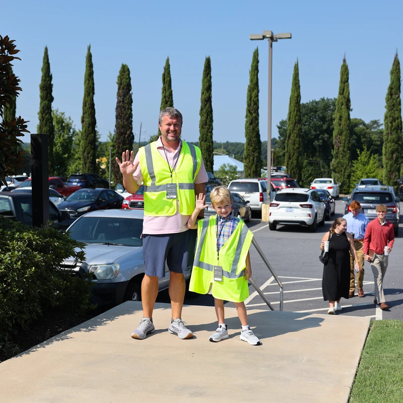 A man and a young boy, both wearing yellow safety vests, smile and wave while standing on a sidewalk next to a busy parking lot on a sunny day.