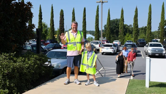A man and a young boy, both wearing yellow safety vests, smile and wave while standing on a sidewalk next to a busy parking lot on a sunny day.