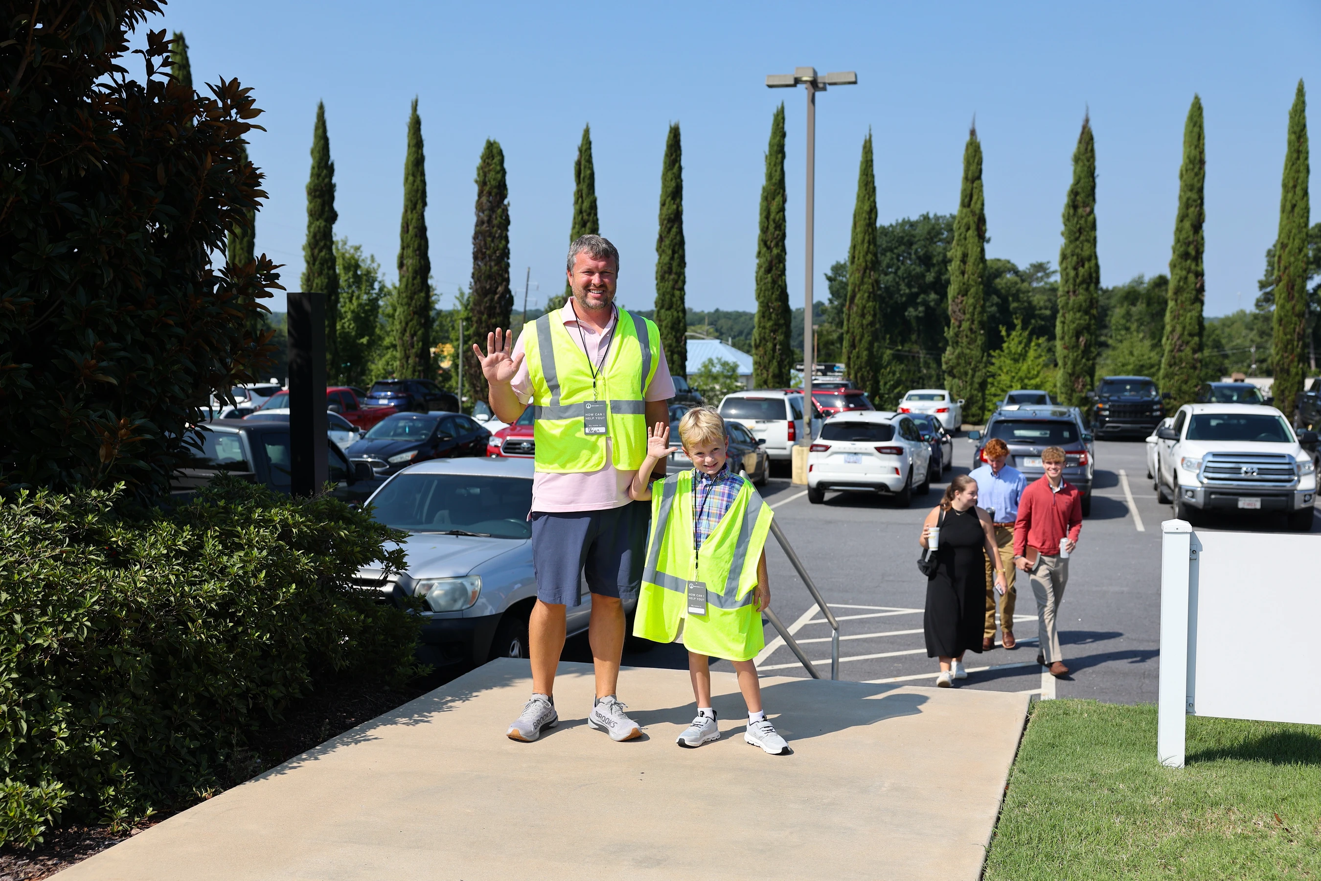 A man and a young boy, both wearing yellow safety vests, smile and wave while standing on a sidewalk next to a busy parking lot on a sunny day.