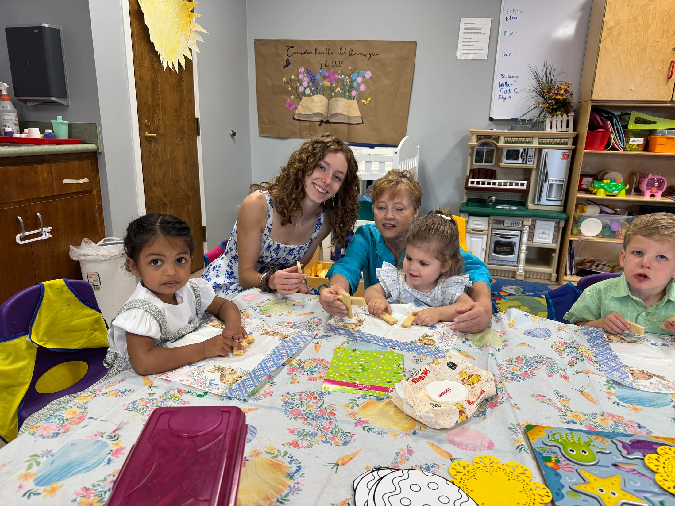 Two women and three young children sit together at a colorful, decorated table in a classroom, smiling and eating wafer cookies during a snack time activity.