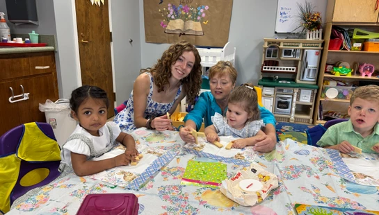 Two women and three young children sit together at a colorful, decorated table in a classroom, smiling and eating wafer cookies during a snack time activity.